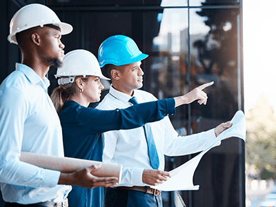 Three professionals in hard hats discussing network cabling project, with one pointing and holding plans, emphasizing collaboration in data center infrastructure development.